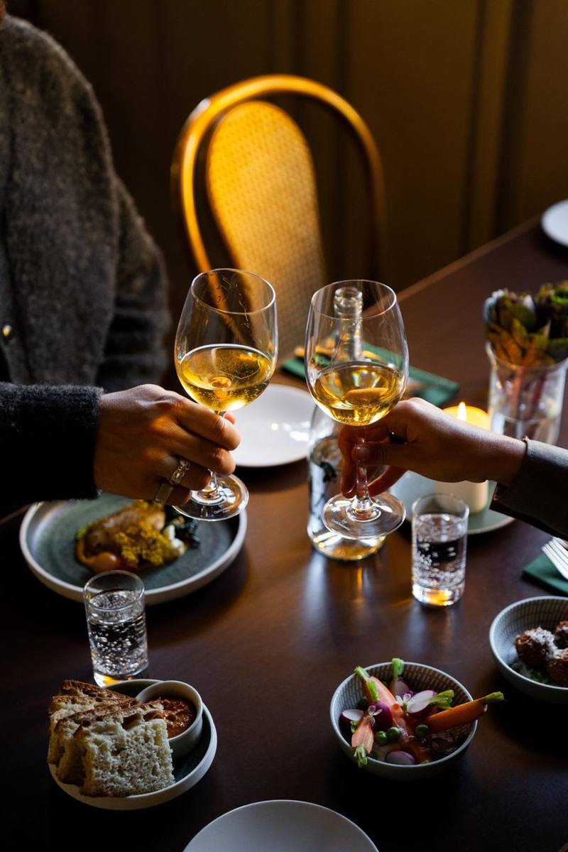 To glas hvidvin skåles over et middagsbord med levende lys på Beghuset